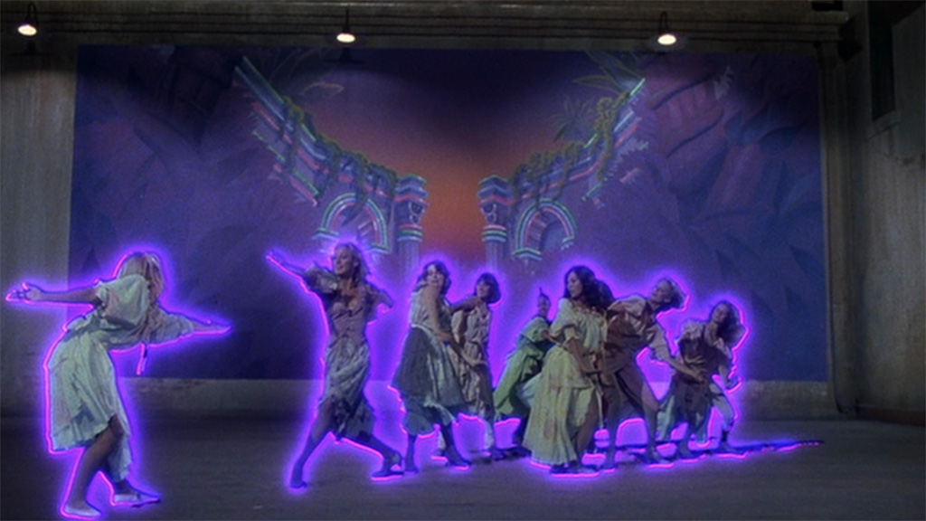 A group of women outlined in purple neon and wearing loose, knee-length dresses dances before a mural showing ancient Greek architecture painted with even more neon.