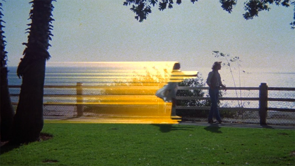 A side shot of a beach scene with grass and a palm tree in the foreground, the ocean in the background. A woman on rollerblades streaks from left to right, trailing yellow neon and reaching for a man walking unsuspecting ahead of her.