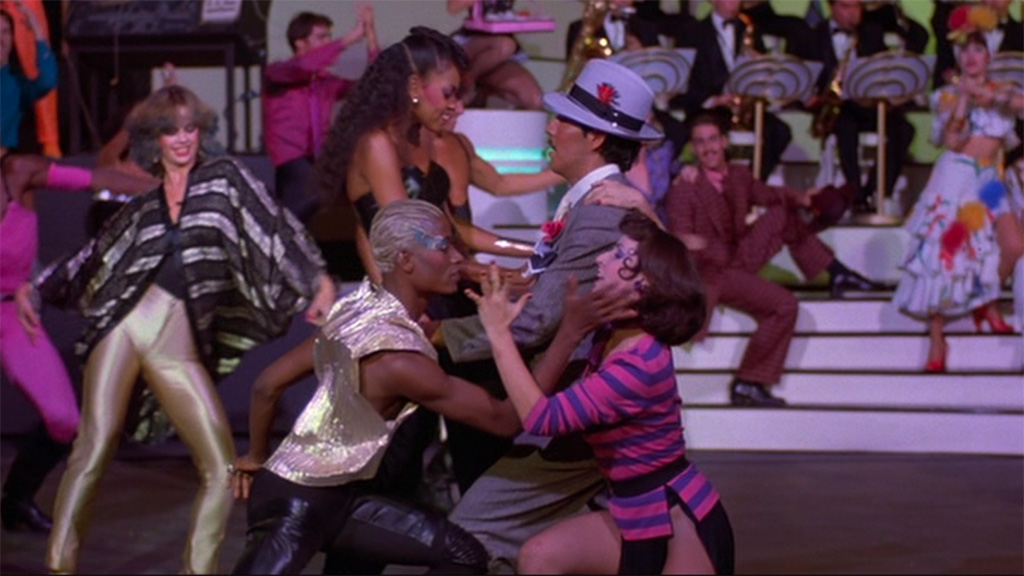 Two pairs of dancers, one dressed in tango garments and the other in new wave spandex, embrace against a backdrop of a big band, a Zoot suit dancer, and a mix of 1980s and 1940s musical groups.