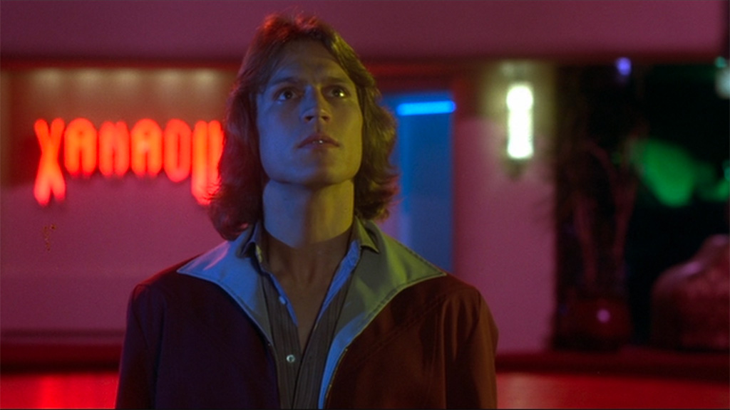 Sonny, a man with feathered hair and a giant, flared color looks up, gazing at where the woman he loves just disappeared in a burst of yellow neon light. Visible in the background over the man's right shoulder is visible a neon red sign, Xanadu.