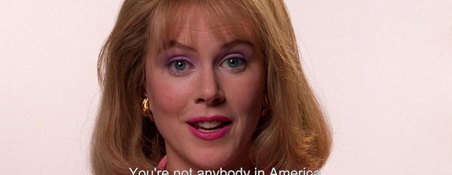 Nicole Kidman with strawberry blonde hair, purple eyeshadow, dark pink lipstick, dark pink jacket, gold earrings, staring into the camera in midspeech with white background. White text, "You're not anybody in America unless you're on TV," fills bottom of image.