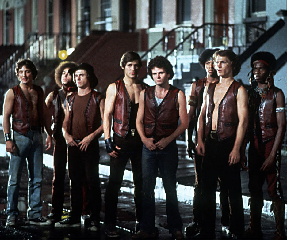 The Warriors, a group of young men, stand on a street in front of brown brick buildings, wearing their signature leather vests