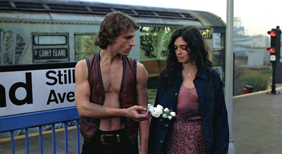 A man, Swan, in a leather vest associated with The Warriors, hands a white corsage to Mercy, a woman in a pink short covered by a black overshirt.