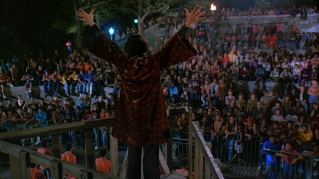 A view from behind, citywide gang orator Cyrus addresses a crowd of hundreds of NYC gang representatives under the cover of night at a park. Dressed in a chocolate brown velour coat and standing on the precipice of a wooden structure, Cyrus's hands are lifted above his head and outstretched in the midst of his call for peace. His audience is rapt, scaffolded on various bits of landscaping, many with arms crossed or hanging over a metal barrier.