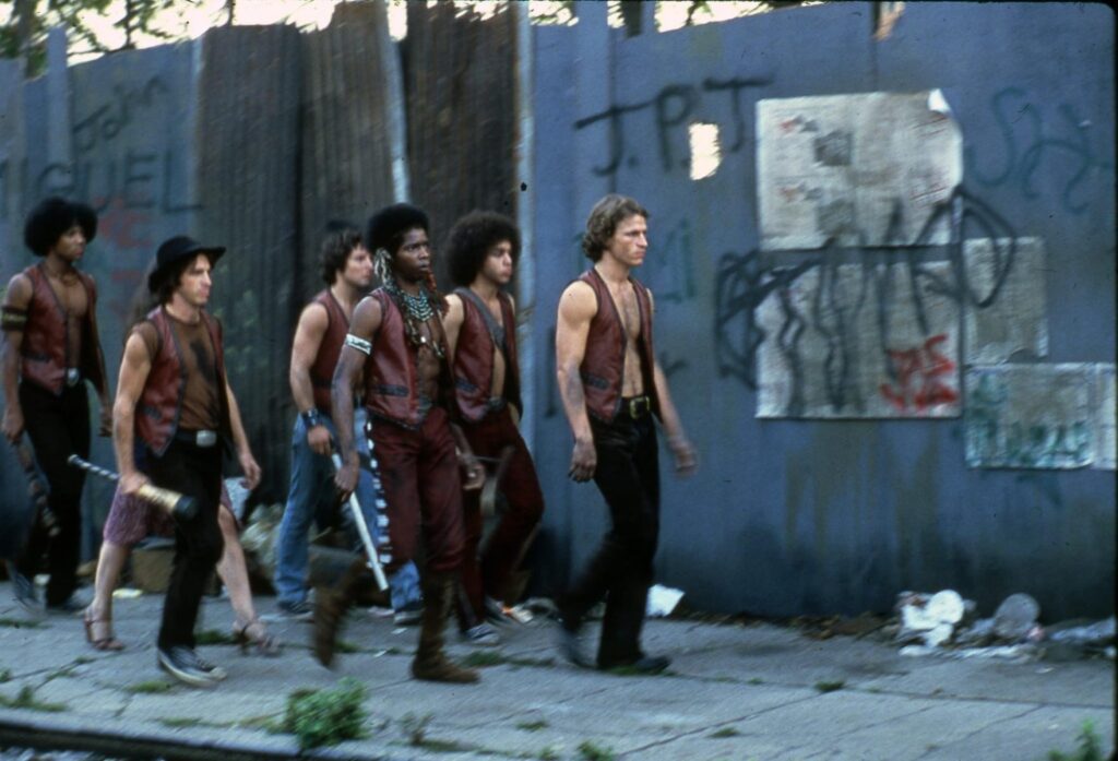 From left to right, the Warriors (plus an interloper) walk a dingy Coney Island sidewalk in front of graffiti-covered solid-wood construction fences in the early morning: Cleon (sporting a large afro), Mercy (the only woman), Cowboy (with a hat to match his name), Vermin, Cochise (adorned with Native American feathers and beads), Rembrandt, and Swan. The gang, whose uniform consists of a brown leather vest and dark pants (only Cowboy wears a shirt underneath), is multiracial, uniformly serious-faced, dirty, and exhausted.