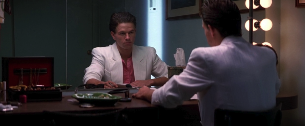 A man with slicked-back hair, a pink undershirt, and a white blazer stares at himself in the mirror.