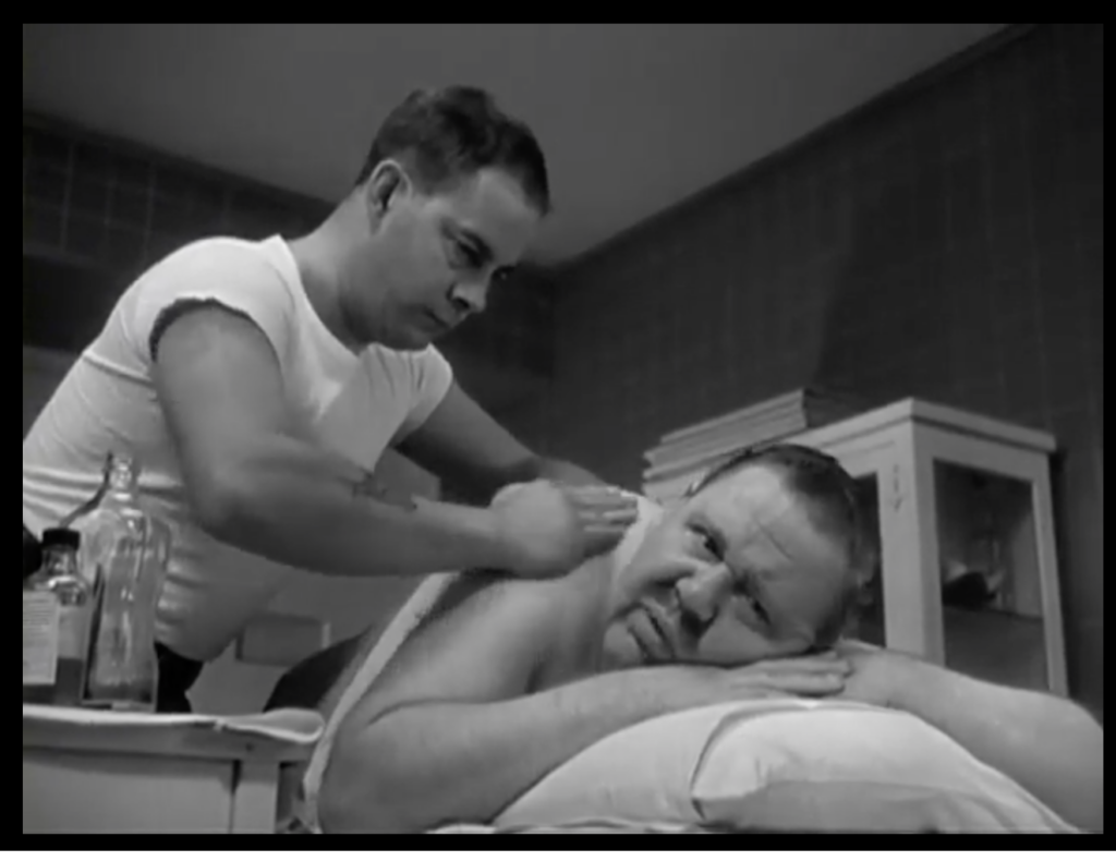 A man lies on a massage table. The masseuse leans over him, rubbing his shoulders and looking intently at the man’s face.