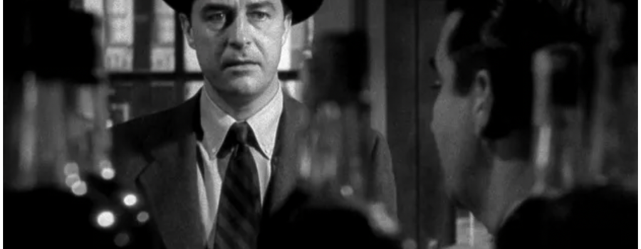 A sweaty man in a suit and hat holding a ten-dollar bill stares at a row of out of focus liquor bottles in the foreground. Image is black and white.