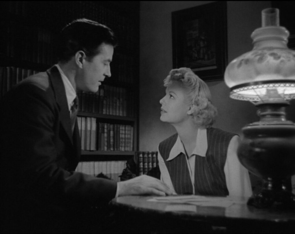 Stephen, wearing a suit and tie, and Carla, wearing a 1940s business suit, lean toward each other across a desk in a study for a kiss