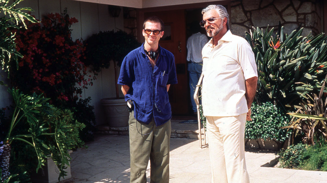 Two men stand in front of a stone-faced house with a variety of flora and foliage covering the exterior. One man is wearing a bkue shirt and kahkis while the other is dressed completely in white. There is someone standing in the back of the photo, facing away from the camera. 