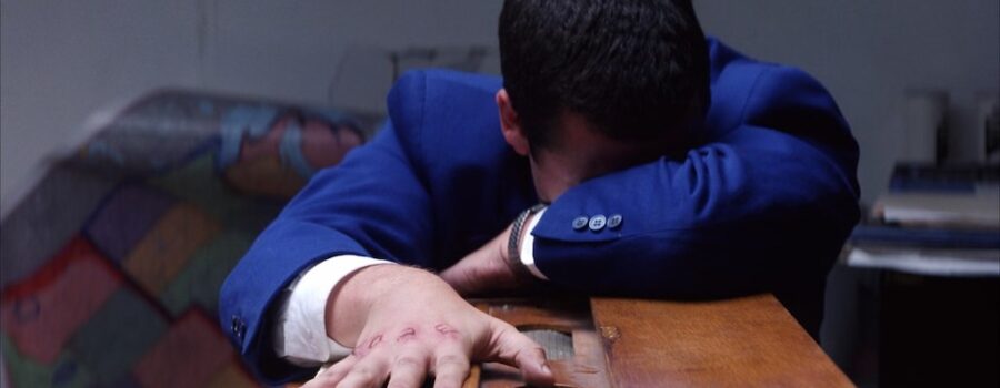 Adam Sandler as Barry Egan leaning over a small harmonium, the word "love" carved into his wounded knuckles, and a torn map hanging off the wall behind him.