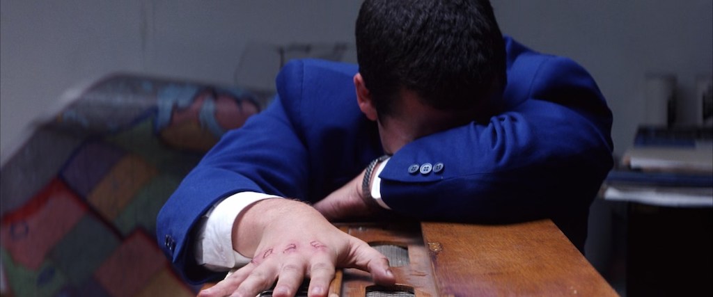 Adam Sandler as Barry Egan leaning over a small harmonium, the word "love" carved into his wounded knuckles, and a torn map hanging off the wall behind him.