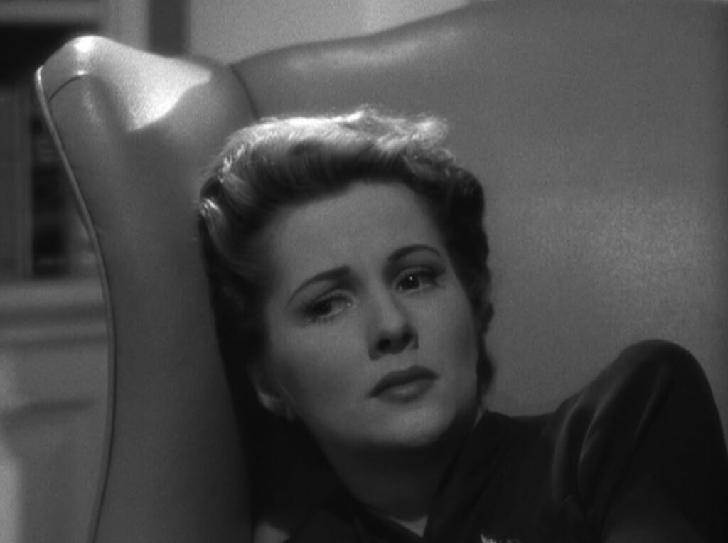 A black-and-white medium close-up of Joan Fontaine as Lina McLaidlaw with her head leaning against the side of a vinyl wingback armchair. A bookshelf and wainscotting are blurred in the background and a light/shadow contrast hits the top of the chair as well as her neck. She is wearing a dark rayon blouse and has a look of deep concern, fright, and/or weariness on her face turned slightly up. her wavy hair is a bit mussed but largely held in a tight updo