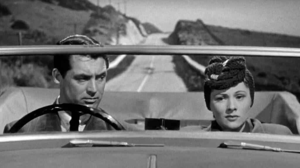 A black-and-white two-shot of Joan Fontaine as Lina McLaidlaw and Cary Grant as Johnny Aysgarth in an English roadster convertible against a hilly, empty two-lane roadway. Grant is in the driver's seat wearing a broad plaid suit with black tie and white shirt, looking at Fontaine on the right in the passenger seat. His eyes are vicious darts, his brow is ominously furrowed, and his dark hair is slicked back. Fontaine's look is one of great trepidation and possible resignment, her eyes aimed straight into a void. Her hair is upturned in a textured fabric wrap.